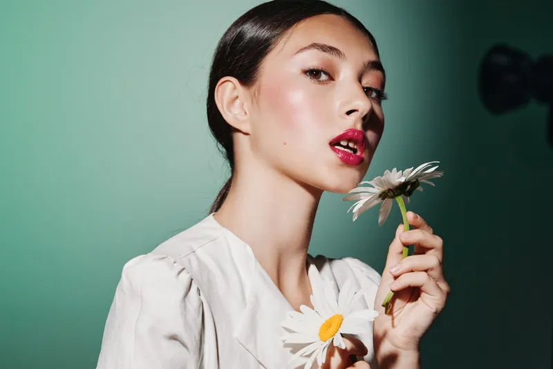 Young model with glossy red lips and flawless skin holds white daisies against a pale green background. Creative fashion beauty portrait showcasing natural floral elegance and makeup art.