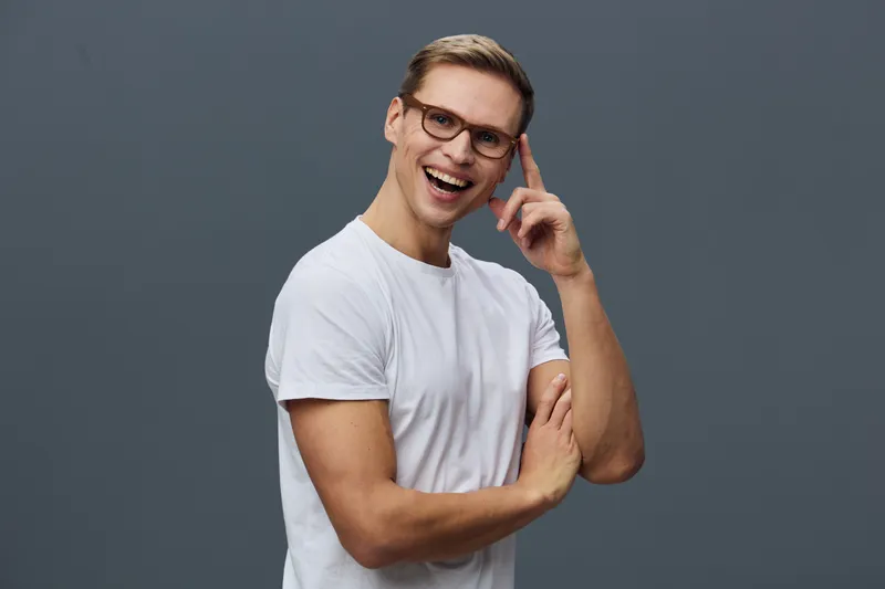 Smiling man with glasses in casual white t-shirt adjusting his glasses and looking at camera on plain gray background. Friendly, cheerful, modern male portrait in studio shot with positive mood. Peopl