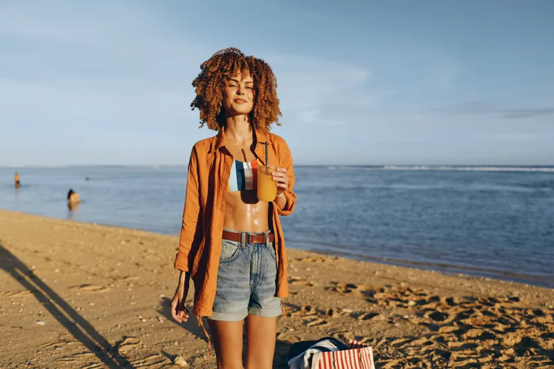 A casual woman on the beach holds mugs of coffee, capturing a sunny, relaxed summer moment. A lifestyle scene focused on travel, leisure, and everyday rituals by the sea. Her denim shorts and orange