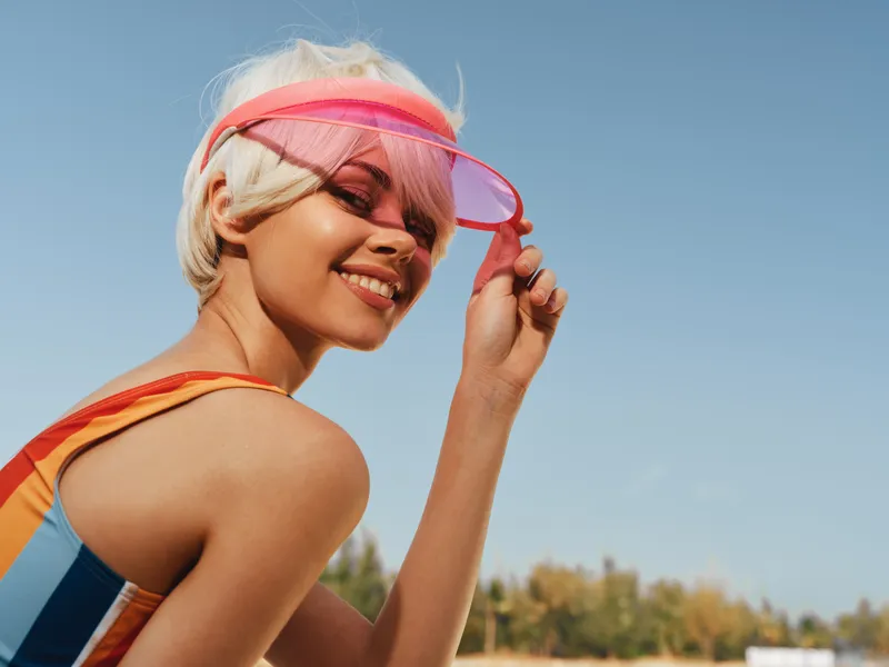 Sunlit portrait of a smiling woman wearing a colorful swimsuit and pink visor, outdoors on a clear day, capturing playful energy, summer fashion, and carefree charm.