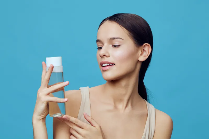 Young woman holding a skincare product, expressing curiosity and happiness against a blue background, showcasing beauty and skincare