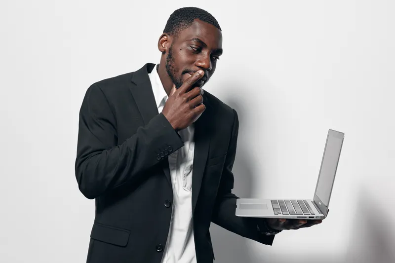 African American man with dark skin in black suit holding laptop and thinking with hand on chin against white background. Business professional, technology and contemplation concept.