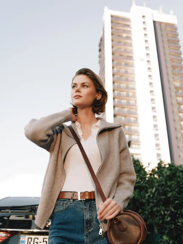 A confident woman stands outdoors in an urban environment, eyes looking ahead with a thoughtful expression. The modern city skyline and warm light add a dynamic, aspirational atmosphere.