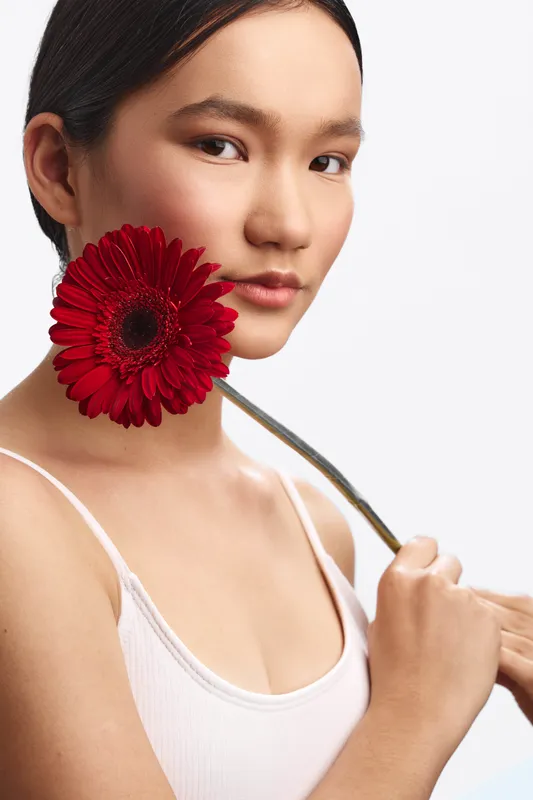 Asian woman posing with a red gerbera flower near her cheek in a clean studio, conveying soft elegance, calm confidence, and subtle glow.
