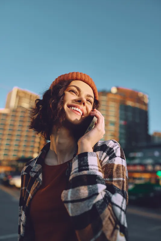 Woman smile phone urban beanie candid portrait in warm golden hour glow, authenticity and candid lifestyle captured with mindful living vibe and emotional storytelling on city streets.