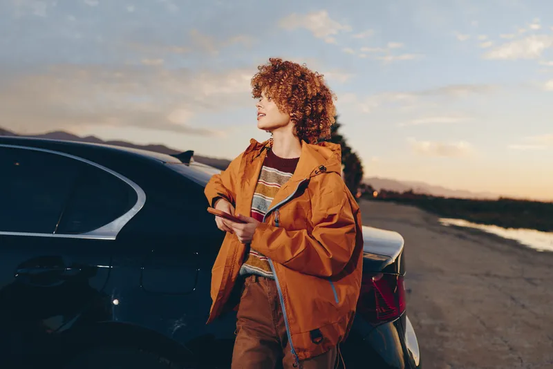 Woman with curly hair in orange jacket stands by car on open road at sunset, relaxed travel lifestyle, outdoor fashion portrait, warm evening sky for solo travel and independence