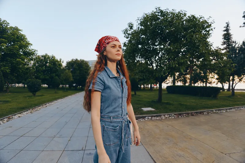 young woman standing outdoors wearing denim jumpsuit and red bandana with trees and pathway in the background during sunset light