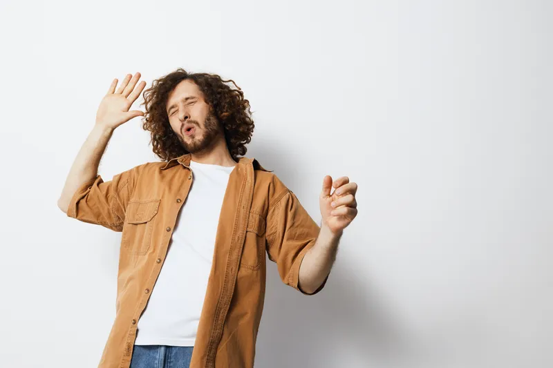 Young man with curly hair in casual clothes joyfully dancing against a light background, expressing happiness and freedom through movement and music