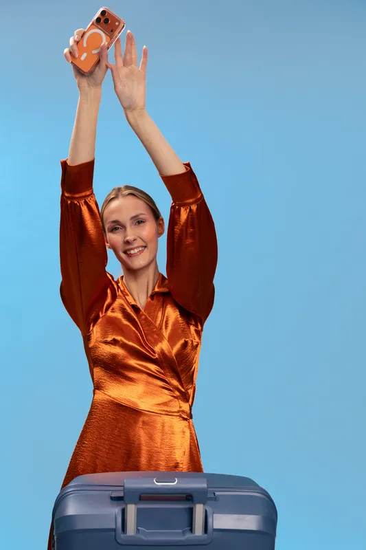 Happy woman in orange dress holds smartphone above head smiling, standing behind blue suitcase. Concept of travel, vacation, technology, and joyful holiday moments in studio with blue background.