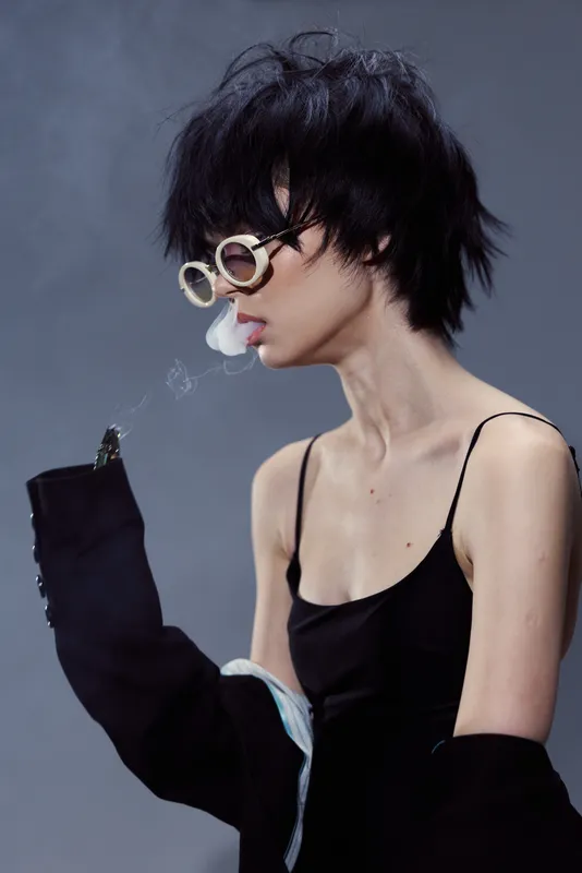 Stylish woman with short hair and sunglasses exhaling smoke wearing black top and gloves