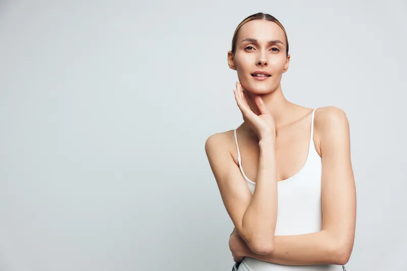 Young slender woman with clear skin and tied hair poses with hand near face wearing white tank top isolated on gray background looking confident and calm.