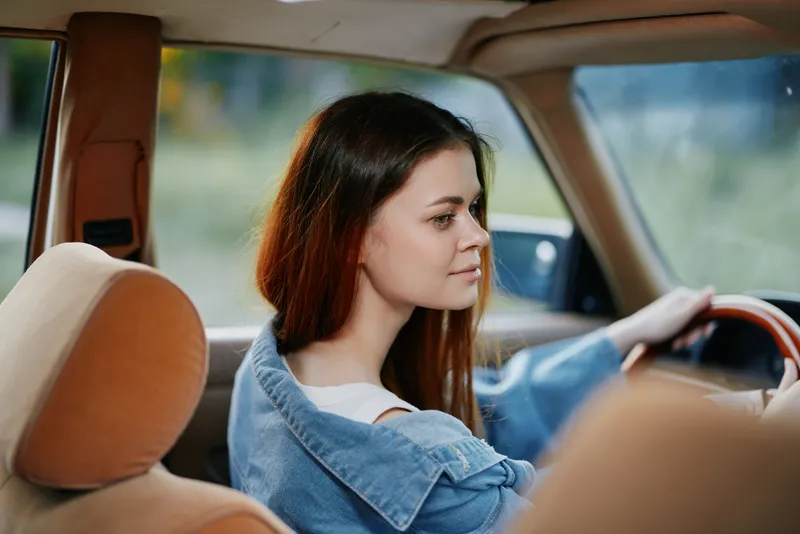 Young woman in a vintage car, smiling while driving, showcasing carefree emotions and a sense of freedom in a natural outdoor setting.