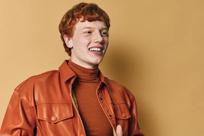 Young man with red hair smiling in brown leather jacket and turtleneck standing against beige background, portrait of cheerful casual style male model happy and confident.