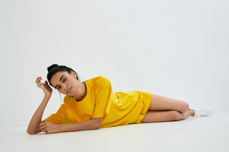 relaxed woman lying on floor wearing bright yellow t-shirt and white sneakers, minimalistic studio background, casual pose and serene mood