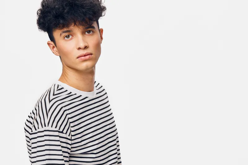 young man with curly hair wearing a striped shirt looks thoughtfully against a white background with soft lighting and minimalistic style