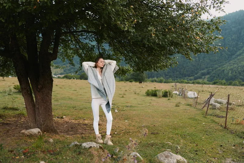 Young woman in a light coat and white pants leans against a tree in a tranquil meadow, enjoying a relaxed moment as distant mountains overlook the scene