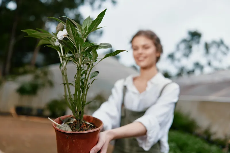 Young woman in casual farming attire cheerfully holding a potted plant against a blurred garden background, symbolizing growth and nurturing nature