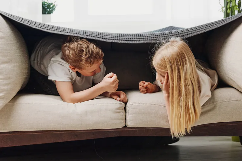 Two children lying under a blanket on a sofa face each other, playing and smiling in a cozy living room with natural light.
