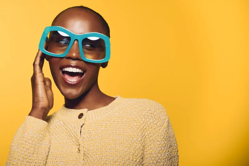 Happy African woman with dark skin wearing large blue glasses and a yellow sweater, smiling joyfully against a bright yellow background. Concept of fashion, style, and happiness.