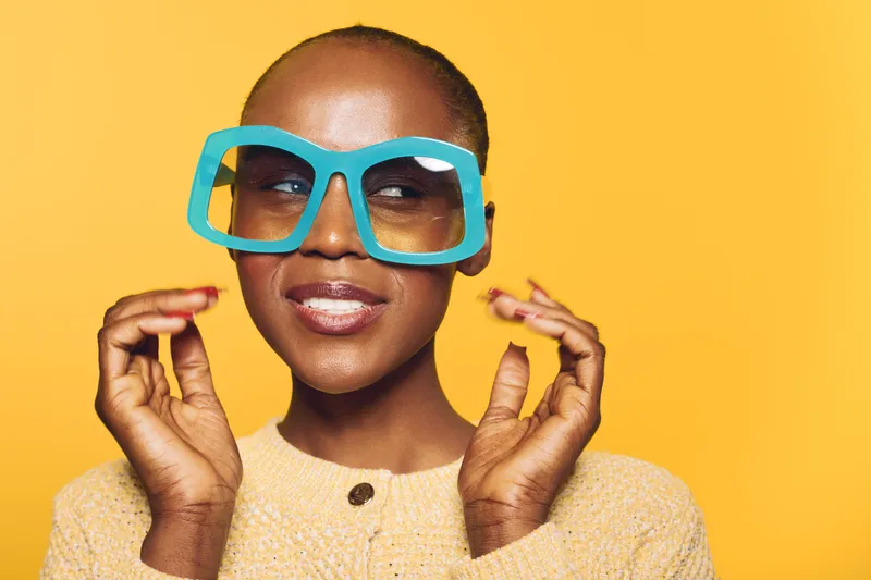 Smiling African woman with dark skin wearing oversized blue glasses and beige sweater poses against vibrant yellow background. Concept of fashion, style, confidence, and beauty.