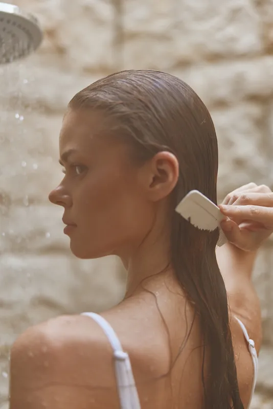 woman combing wet hair in shower with neutral stone background, natural light, intimate skincare moment, fresh and calm atmosphere