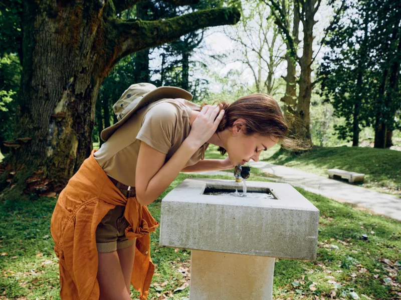 Woman drinking water from a concrete fountain in a park, wearing backpack and shorts, leaning forward to sip from the spout, casual summer outfit and green nature background.