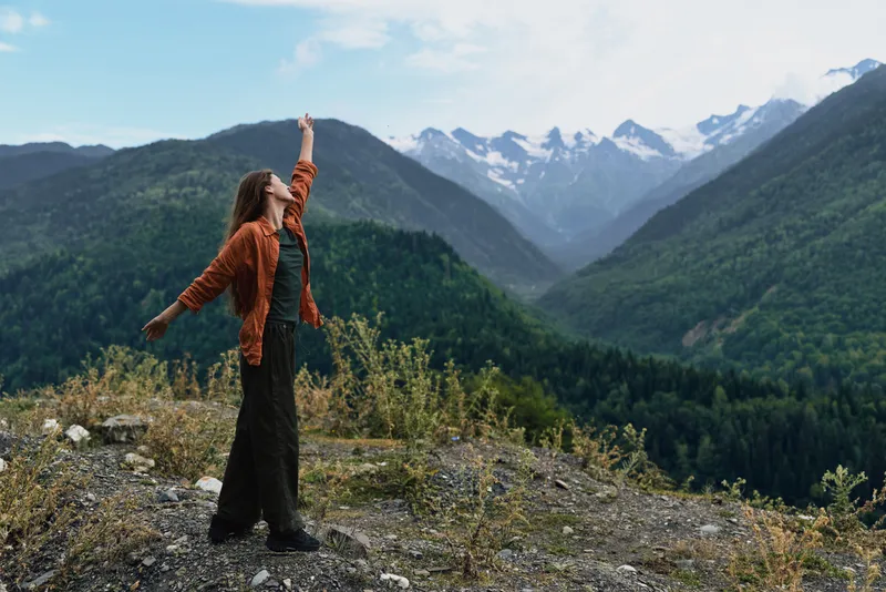 Woman stands on a rocky overlook with raised arm amid vast mountains and valleys, radiating freedom and adventure.
