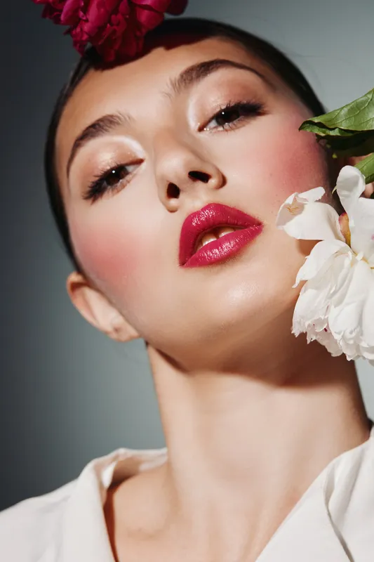 Young woman with glossy red lips and highlighted cheeks posing with floral accessories on her head and neck, beauty fashion creative studio portrait with dramatic lighting.