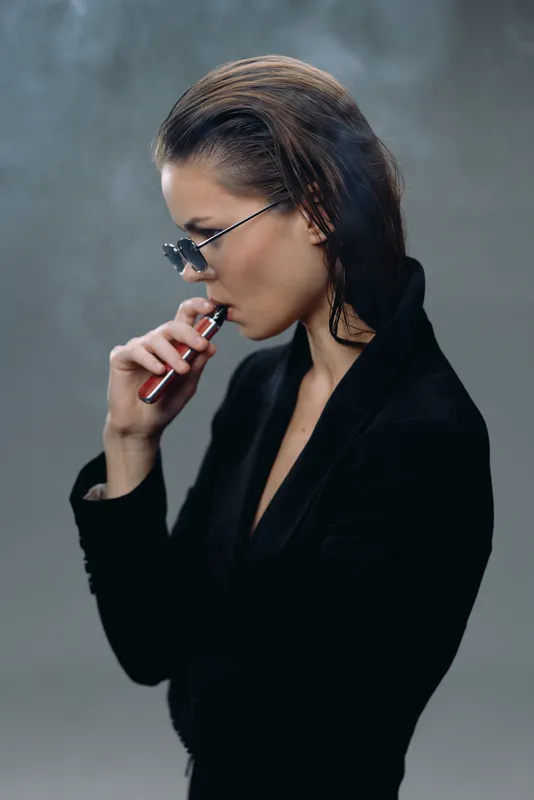 Young woman vaping with sunglasses in a dark smoky atmosphere wearing a black blazer and slicked-back hair.