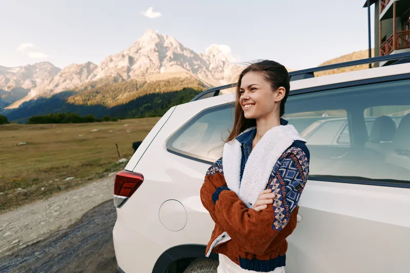 Woman leaning on white car by mountain road, smile on her face while travel outdoors, scenic mountains and landscape, casual jacket and relaxed posture during a leisure trip.