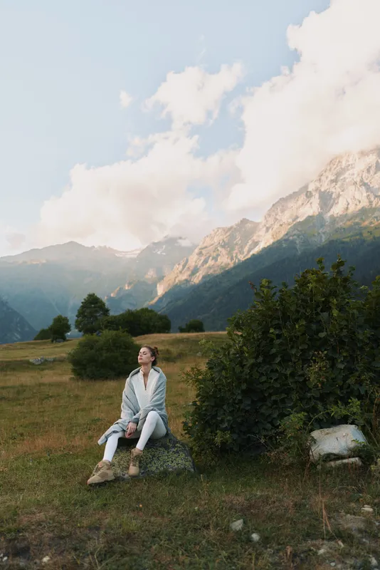 Young woman in a light denim jacket and jeans sits on a rock in a green meadow, with distant alpine mountains and a bright sky forming a dramatic natural backdrop