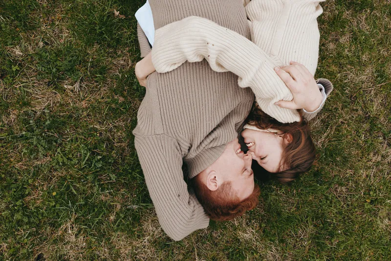 Couple outdoors, lying on grass, embracing each other in cozy sweaters, warm natural tones, tender moment, soft breeze, candid lifestyle, peaceful mood, intimate connection, quiet romance