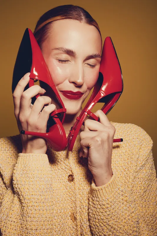 Young woman with closed eyes holds red high heels near face, smiling softly. Concept of fashion, style, beauty, and joyful emotions in studio lighting.