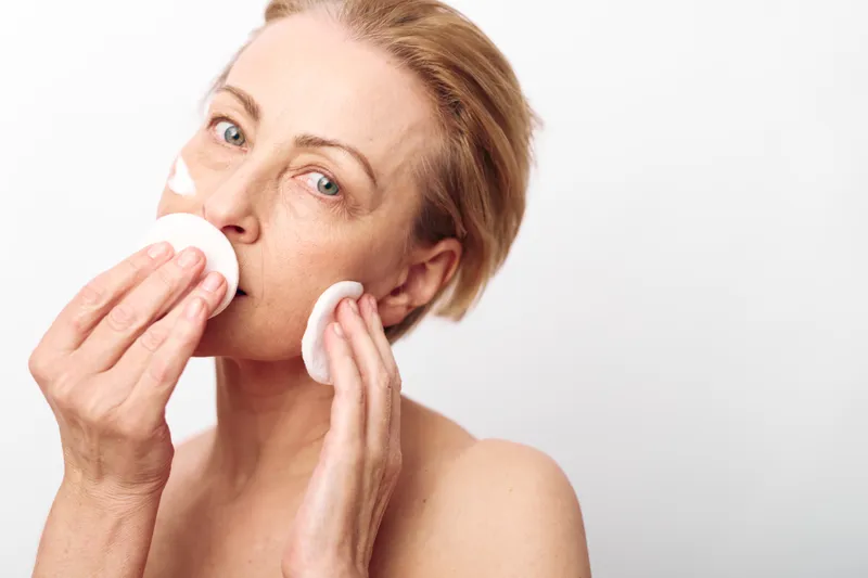 Mature woman with light skin removes facial cream using cotton pads on white background. Skincare routine concept for natural beauty, self-care, and healthy skin at home in soft daylight.