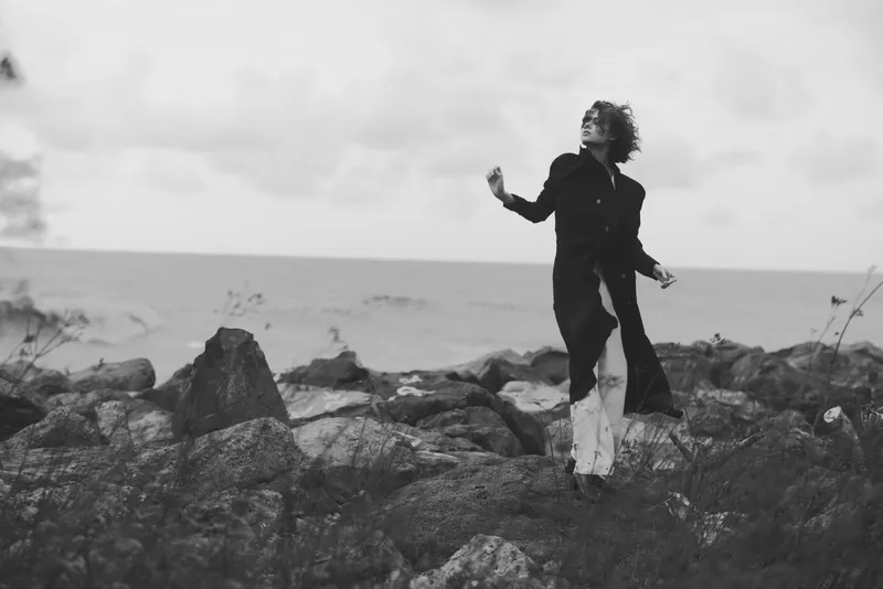 Black and white scene of a dancer on a rocky shoreline mid motion with an expressive pose wind in hair waves and horizon behind artistic movement moody atmosphere and performance energy
