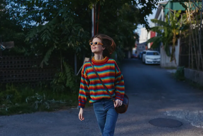 Young woman in a colorful striped sweater and jeans, smiling and walking down a quiet street wearing sunglasses and a crossbody bag, relaxed urban lifestyle travel vibe.