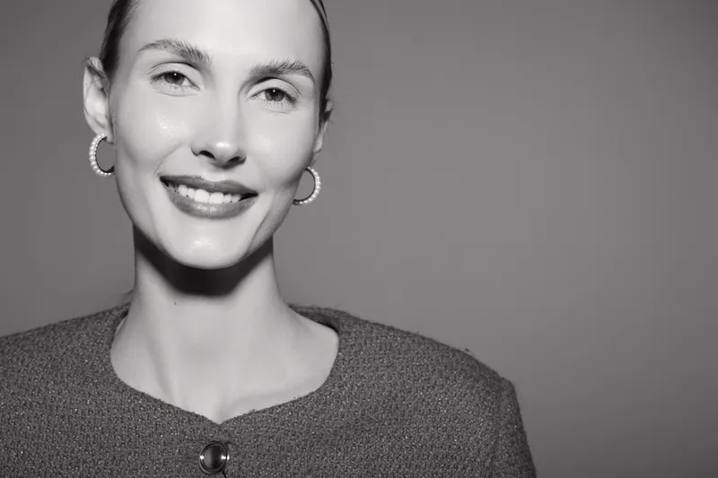 Smiling Caucasian woman with light skin and slicked hair looks at camera wearing hoop earrings and textured jacket. Studio portrait with neutral background.