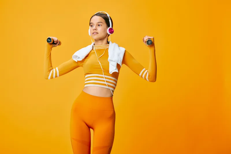 Fitness woman in bright orange athletic wear lifts dumbbells with a towel and headphones on an orange backdrop, showcasing strength, energy and focus during a dynamic workout.