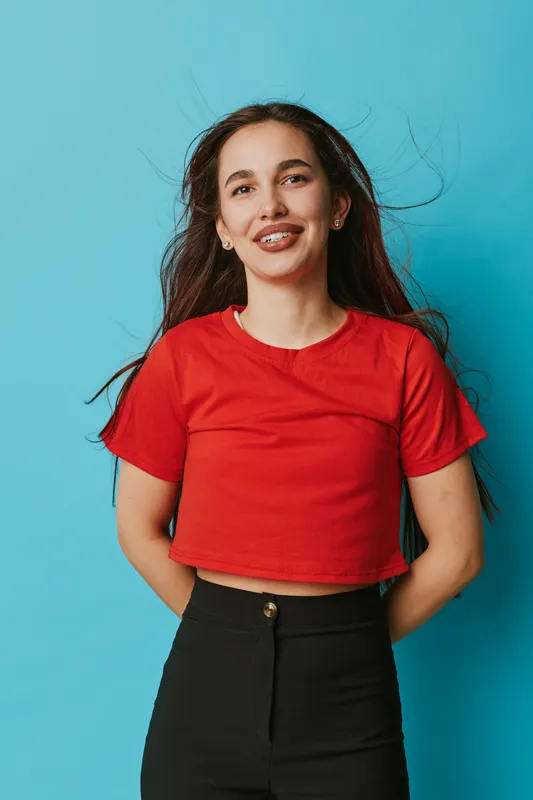 Young woman with long hair wearing red crop top and black pants stands smiling with hands behind back against blue background. Casual style and confident pose in studio.