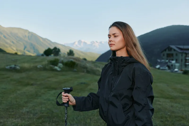 A focused hiker stands outdoors in a rugged landscape, holding a trekking pole with a calm expression. She wears a dark rain jacket, ready for a mountain trek.