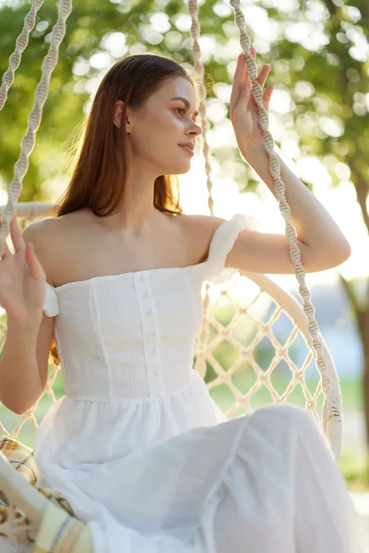 Young woman in a white dress enjoying a serene moment on a swing in a sunny outdoor setting, embodying tranquility and natural beauty