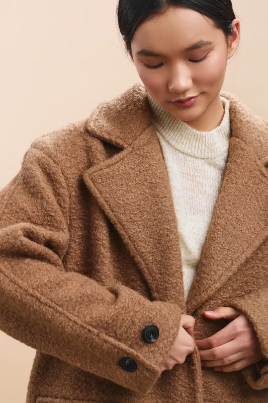asian woman in a cozy beige coat adjusting the front, calm expression, soft studio backdrop, warm tones convey elegance and style