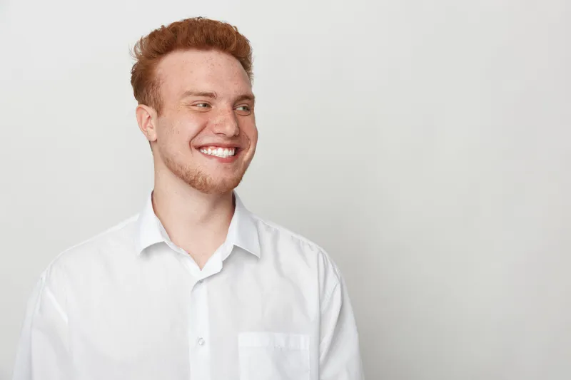 Smiling young man with red hair wearing white shirt looks away on light background. Casual portrait of happy male model with natural expression and clean studio lighting.