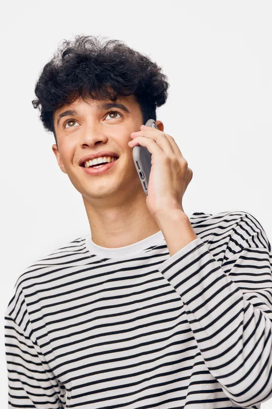 young man smiling talking on smartphone wearing striped shirt with curly hair against white background in casual style, positive expression, communication concept