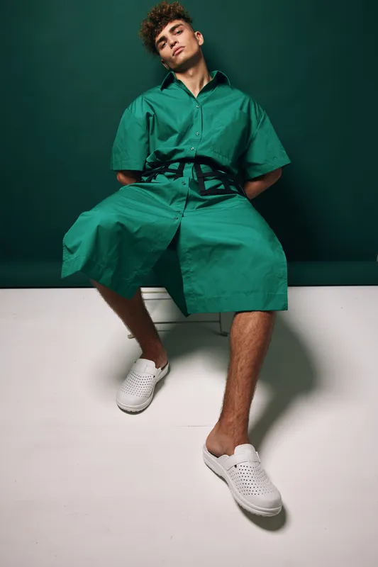 Young man in green oversized shirt and shorts sitting on white bench against dark green background with casual white slip-on shoes and relaxed pose in studio lighting.