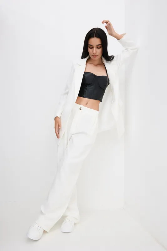 Stylish woman wearing white wide pants and jacket with black crop top striking a chic pose against a minimal white background, conveying modern fashion and confidence.