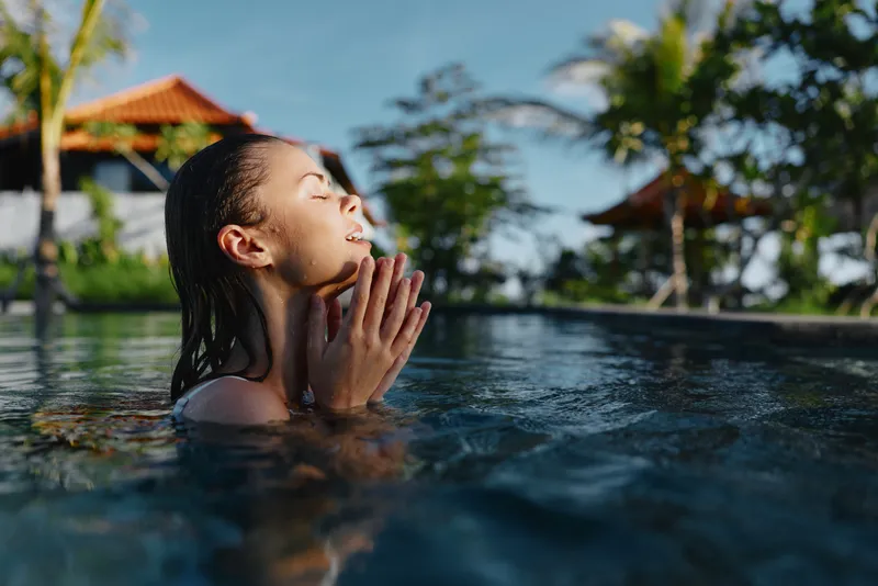 A woman relaxes in a tropical pool, smiling with hands pressed together in a peaceful greeting, showcasing leisure lifestyle and wellness at a sunny resort setting during vacation.