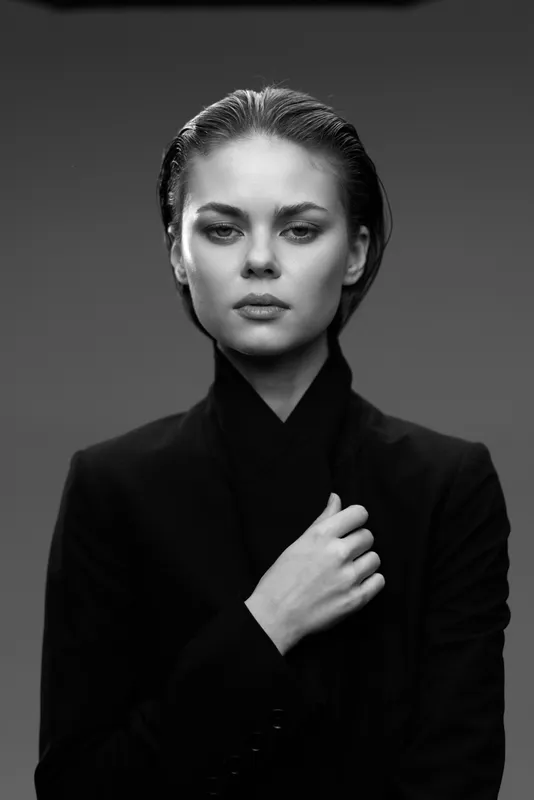 Black and white portrait of a serious woman with slicked-back hair wearing a black suit on a plain background