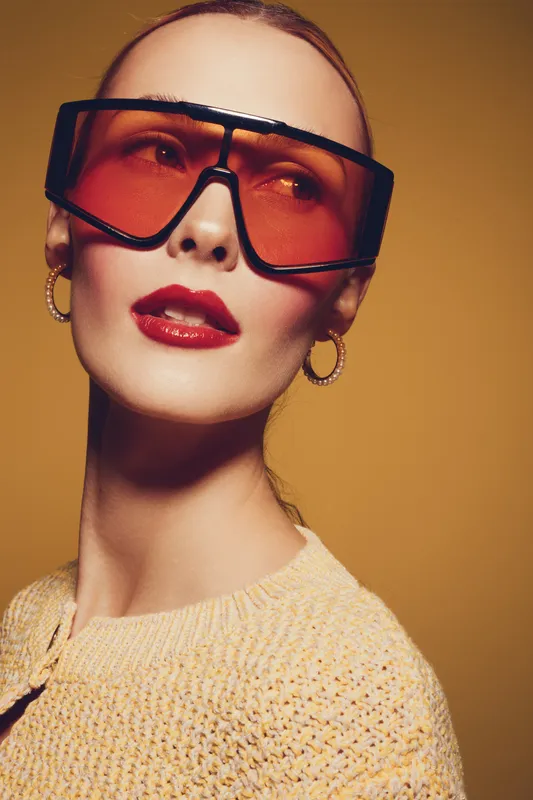 Fashionable woman with oversized orange sunglasses and red lipstick poses against warm yellow background. Concept of modern style, beauty, and confident attitude in studio lighting.