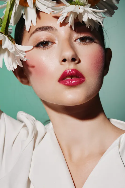 Close-up of female model with red glossy lips and pink blush wearing white petals on head. Studio portrait on green background. Concept of beauty, fashion, and creative makeup.